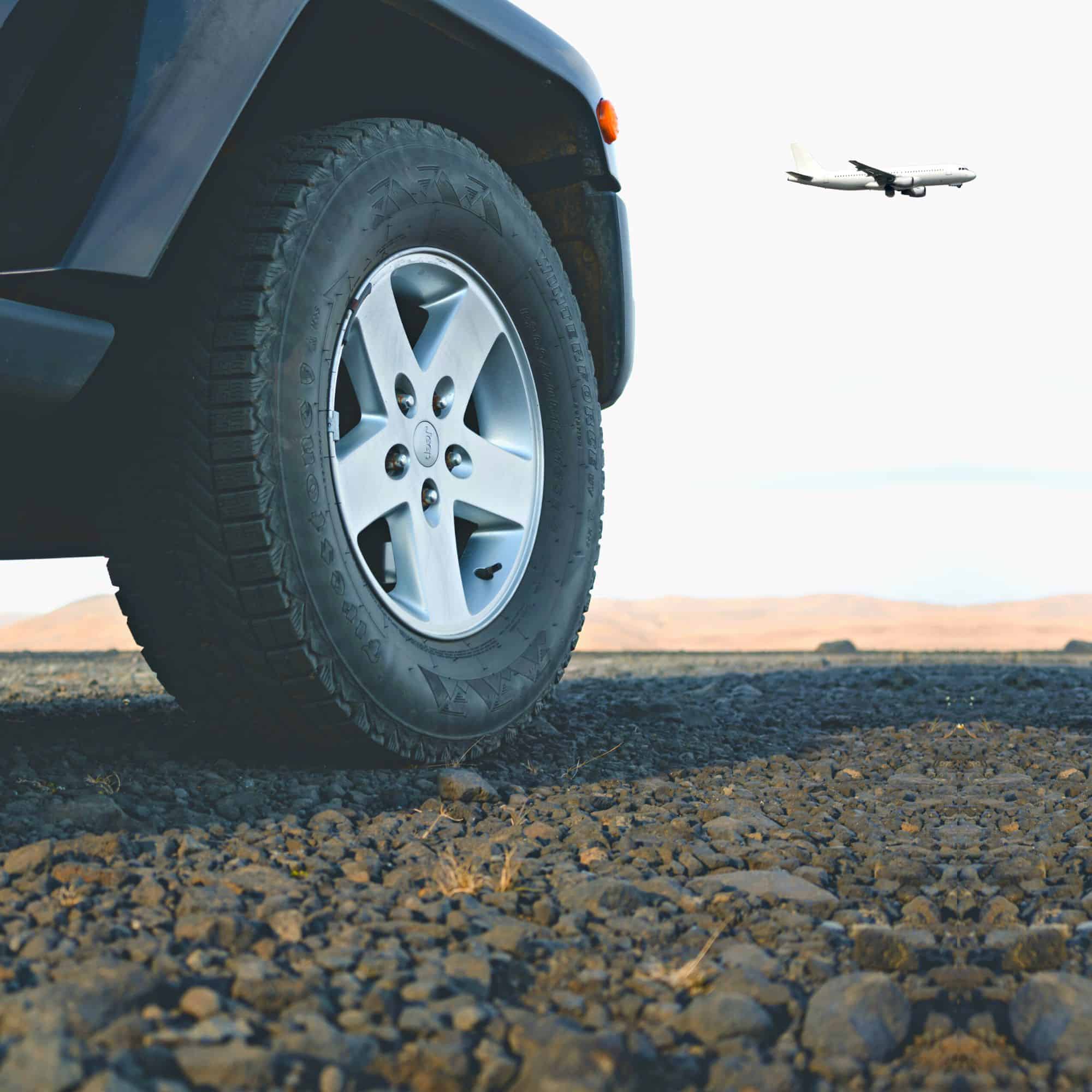 A low angled photo of a plane flying through the sky in the background. A graveled road and a stationary car tyre is in the foreground.