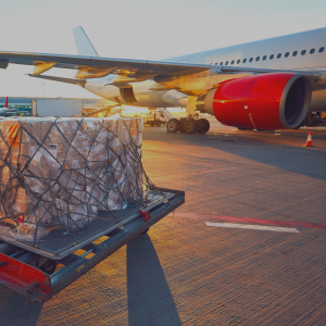 A palette of goods is standing on the runway in the foreground of the image. In the bnackground a plane is waiting, with its cargo door open. The sun is shining.