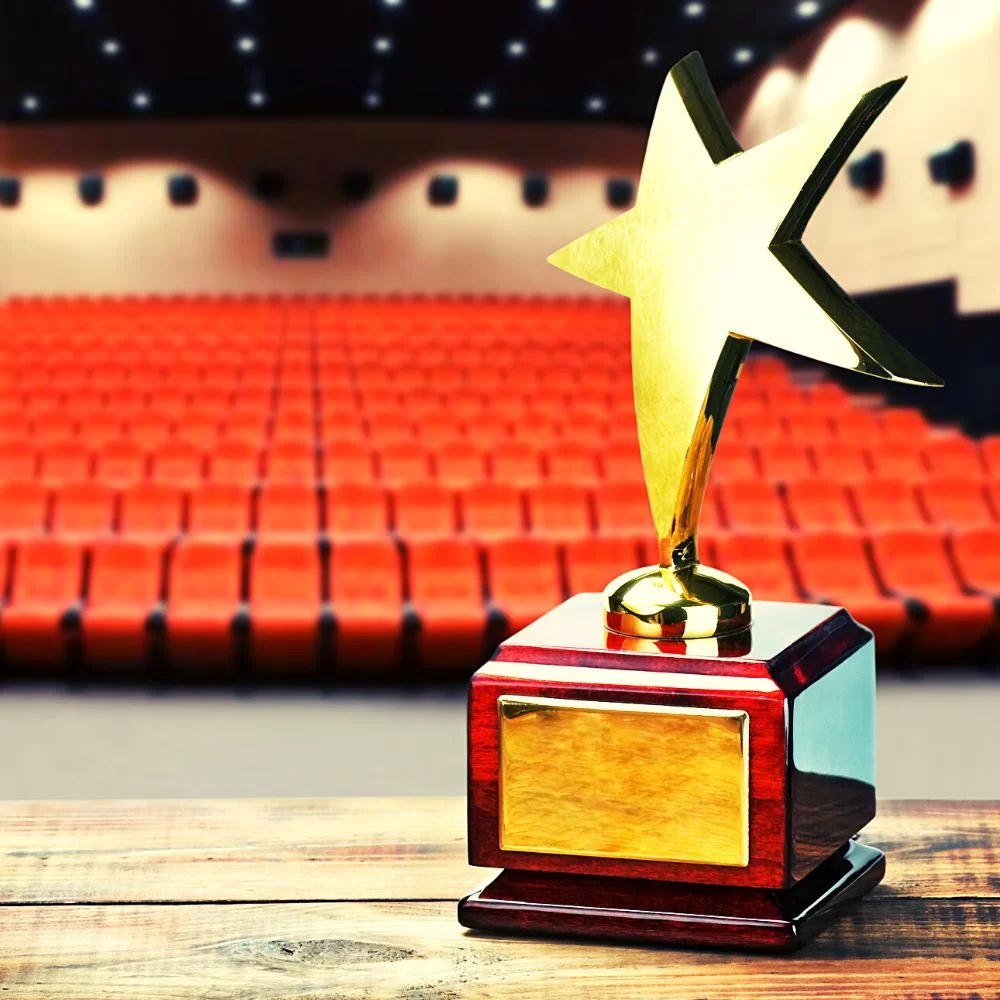 An gold award in the shape of a star with a polished wooden base and blank plaque sits on a tabletop in the foreground of the image. The background is blurred and shows rows of red seats in an empty auditorium of a theatre