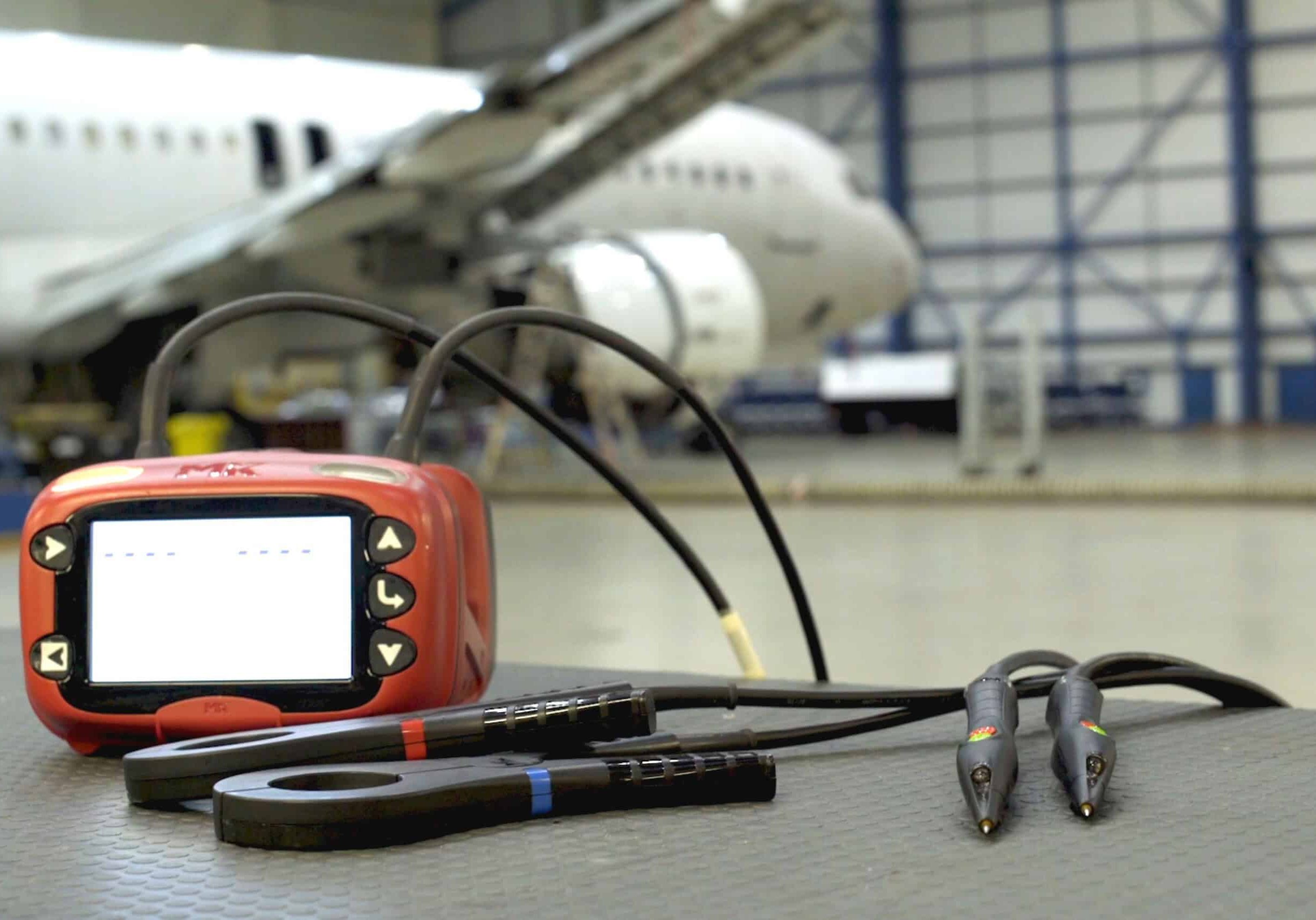ExLRT tool is positioned on a table in the foreground, with the couplers and probes in front of it. In the background is a plane in a maintenance hangar.